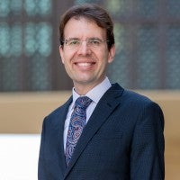 A man wearing glasses and a dark suit with a patterned tie is smiling at the camera. The background is blurred with geometric patterns and muted colors.
