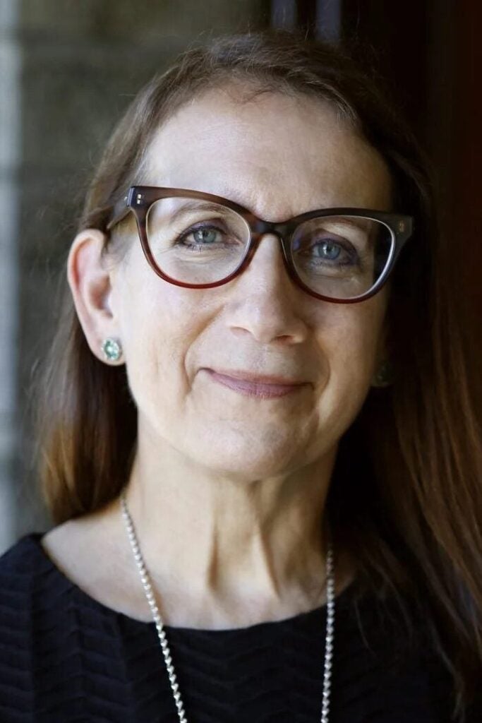 A woman with long brown hair and glasses smiles gently. She is wearing a black top, green earrings, and a silver necklace, standing in front of a softly blurred background.