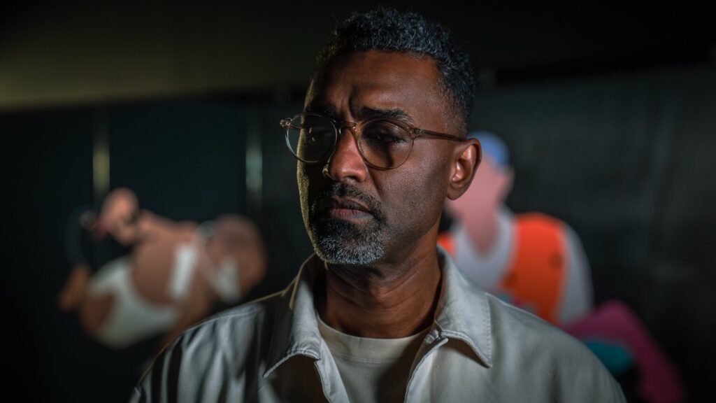 A man with short curly hair, glasses, and a gray goatee stands in focus, wearing a light-colored jacket. The background is blurred with indistinct shapes and colorful figures.