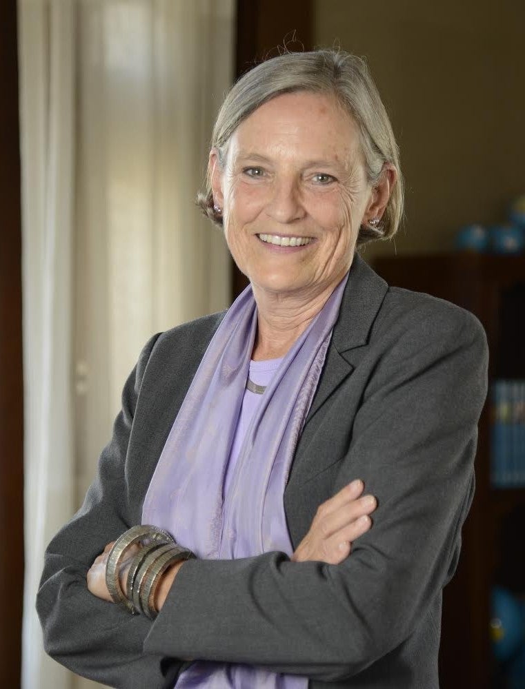 An older woman with short gray hair, wearing a gray blazer, lavender scarf, and bracelets, stands smiling with arms crossed in an indoor setting with shelves and curtains in the background.