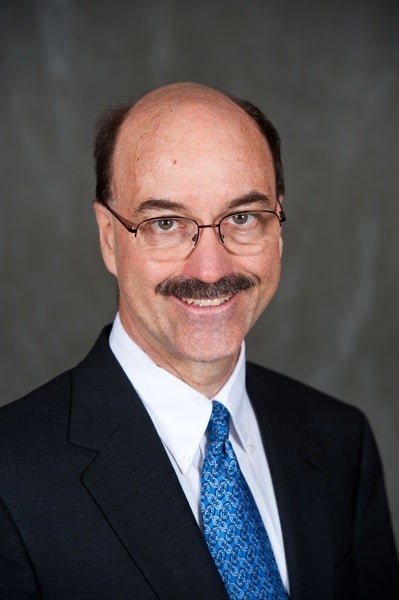 A middle-aged man with glasses, a mustache, and thinning hair, wearing a dark suit, white shirt, and blue patterned tie, smiling at the camera against a gray background.