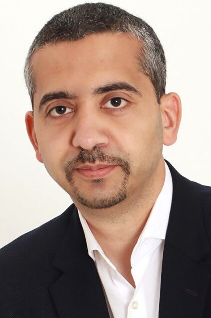 A man with short, dark hair and a trimmed goatee, wearing a white shirt and a dark blazer, is looking at the camera against a plain white background.