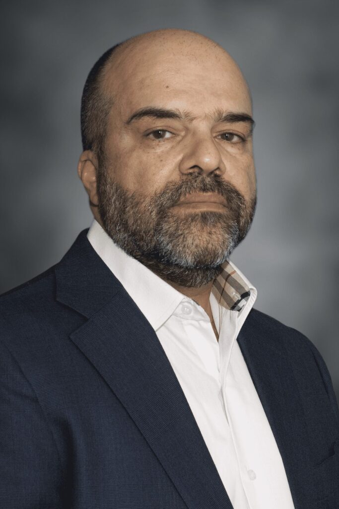 A middle-aged man with a shaved head and beard wearing a white shirt and dark blue blazer poses for a formal portrait against a gray background.