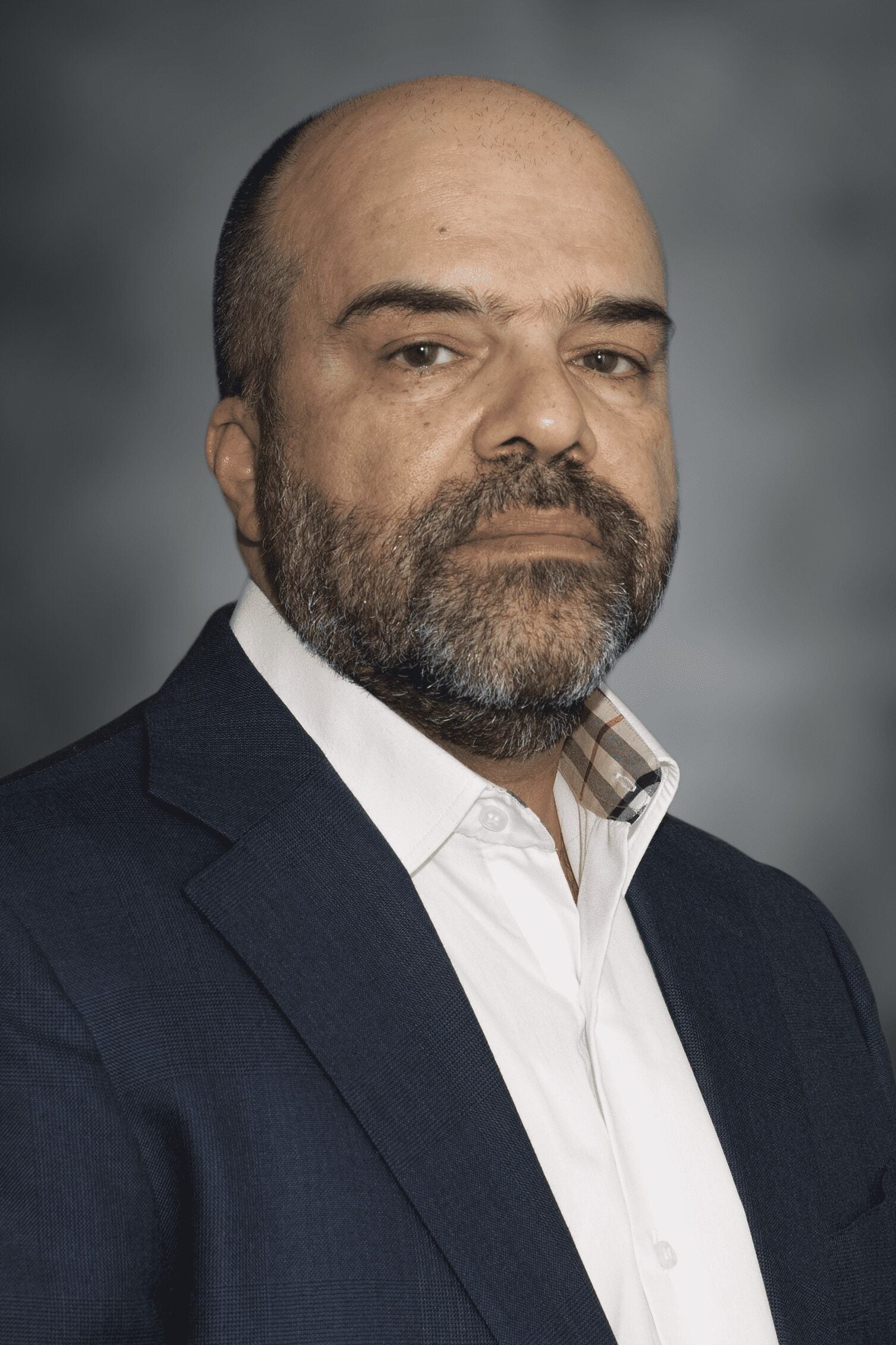 A middle-aged man with a shaved head and beard wearing a white shirt and dark blue blazer poses for a formal portrait against a gray background.
