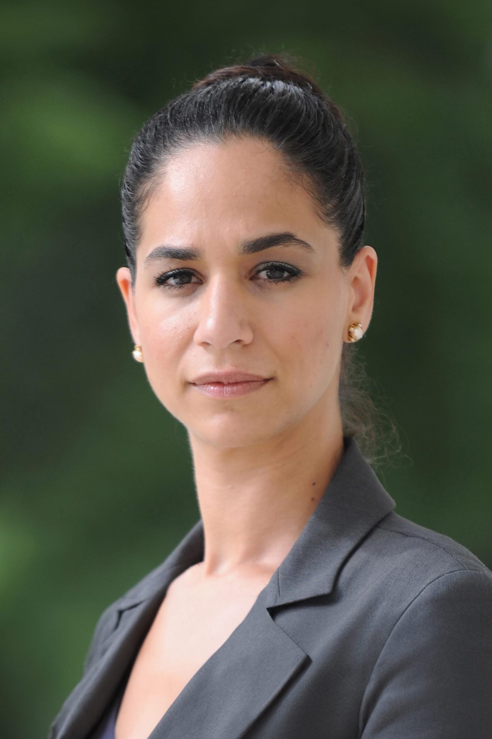 A woman with dark hair pulled back, wearing a gray blazer and gold stud earrings, stands outdoors against a blurred green background, looking confidently at the camera.