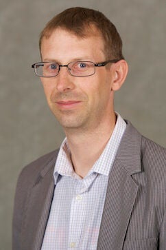 A man with short light brown hair, wearing glasses, a light plaid shirt, and a gray blazer, poses against a plain gray background.