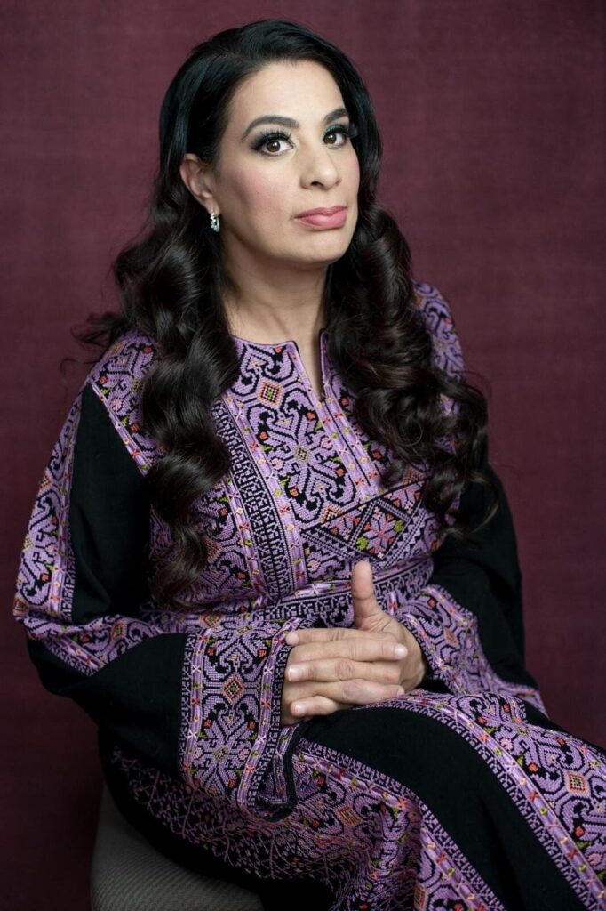 A woman with long dark hair and light makeup sits against a maroon background, wearing a traditional embroidered dress in purple and black. Her hands are folded in her lap, and she looks slightly to the side.