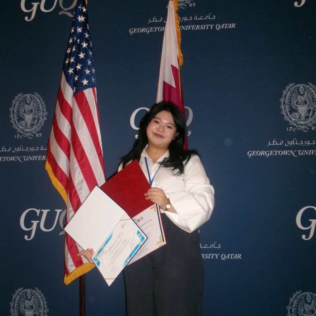 Student with brown hair and white shirt stands with her certificates from GPS in front of Qatar and USA flags with GU-Q branded blue background