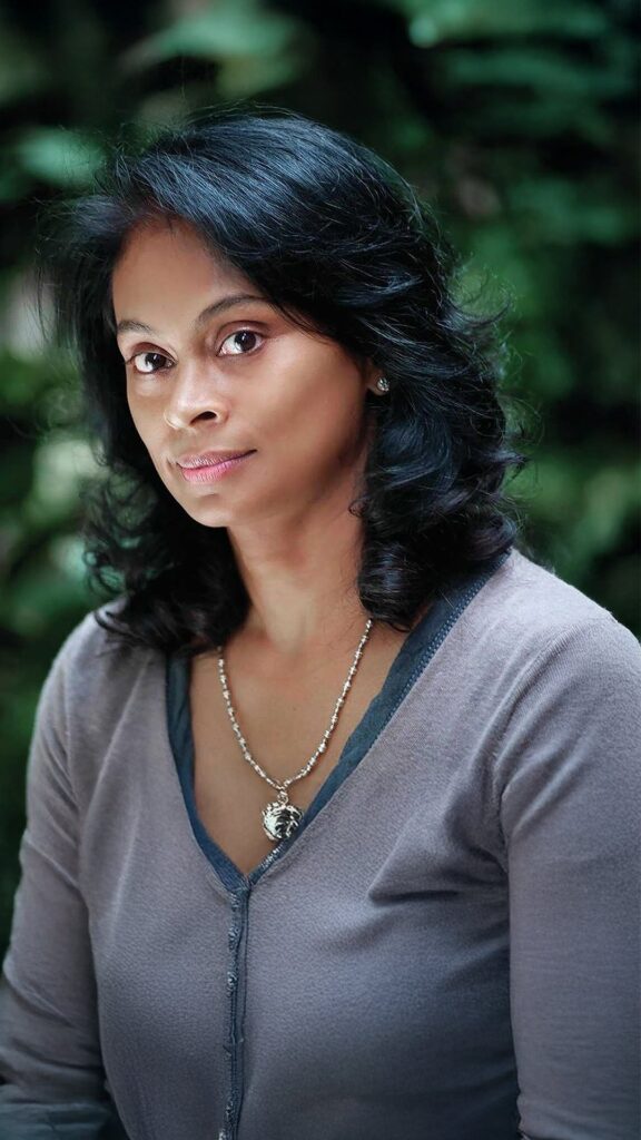 A woman with medium-length black hair and a serious expression wears a gray top and a silver necklace, standing in front of a blurred green background.