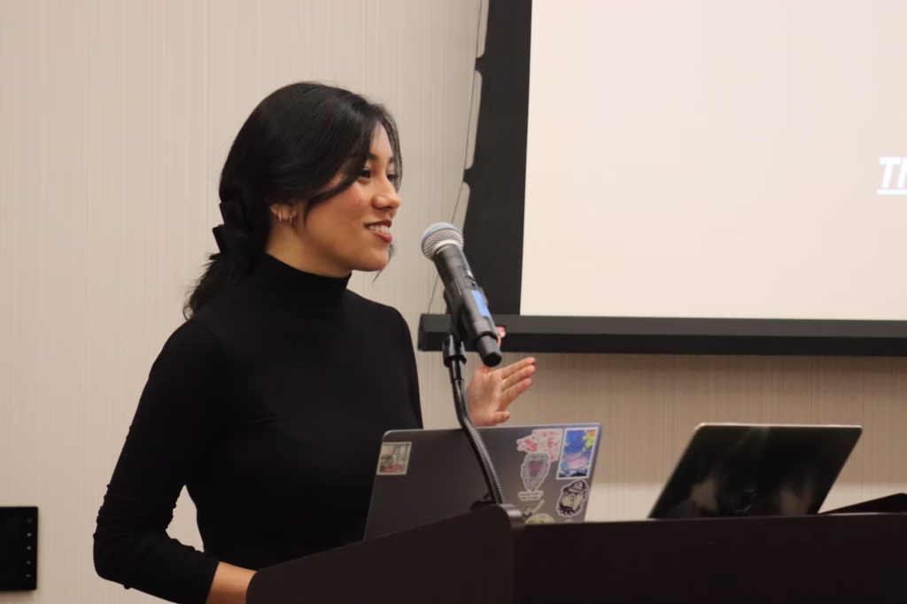 A woman with long dark hair, wearing a black turtleneck, speaks at a podium with a microphone. Two laptops, one covered in stickers, are on the podium. A projection screen is visible in the background.