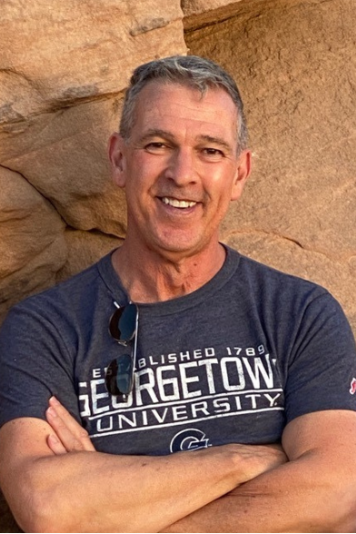 A man with short gray hair smiles with arms crossed, wearing a navy blue Georgetown University T-shirt and sunglasses hanging from the collar, standing in front of large tan rocks.