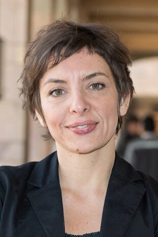 A woman with short, tousled brown hair and light eyes is smiling slightly. She is wearing a black blazer and standing in an outdoor setting with blurred people in the background.
