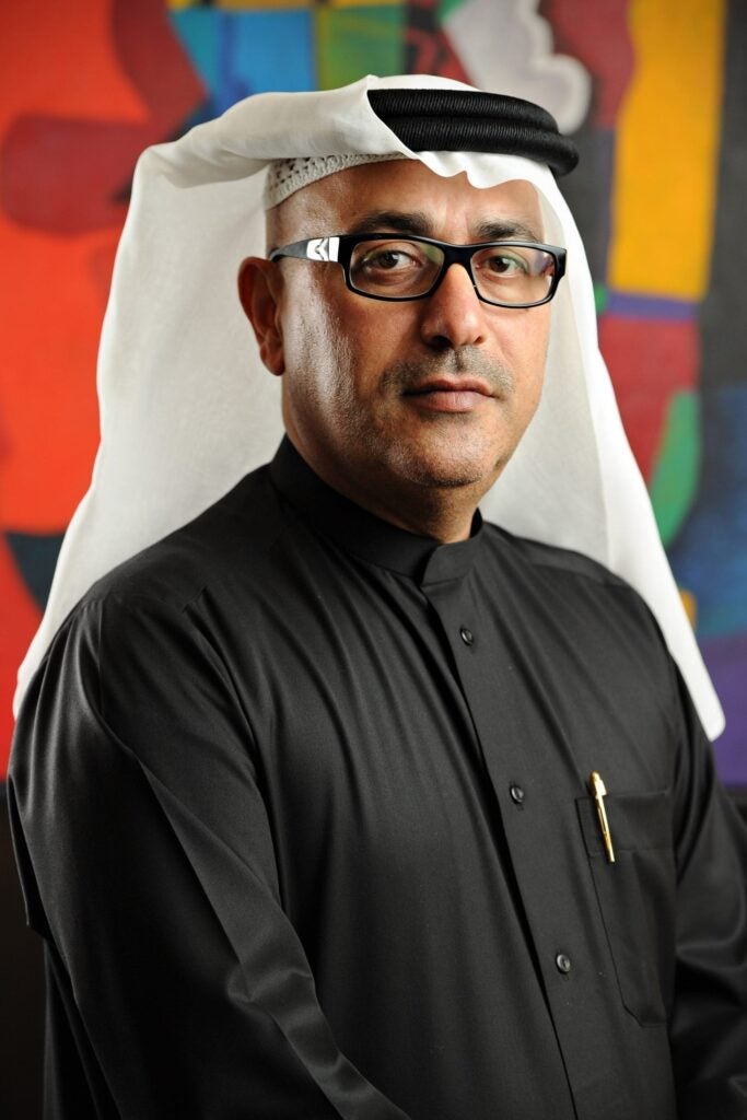 A man wearing glasses, a white keffiyeh, and a black thobe stands in front of a colorful abstract background. He has a neutral expression and a pen in his shirt pocket.