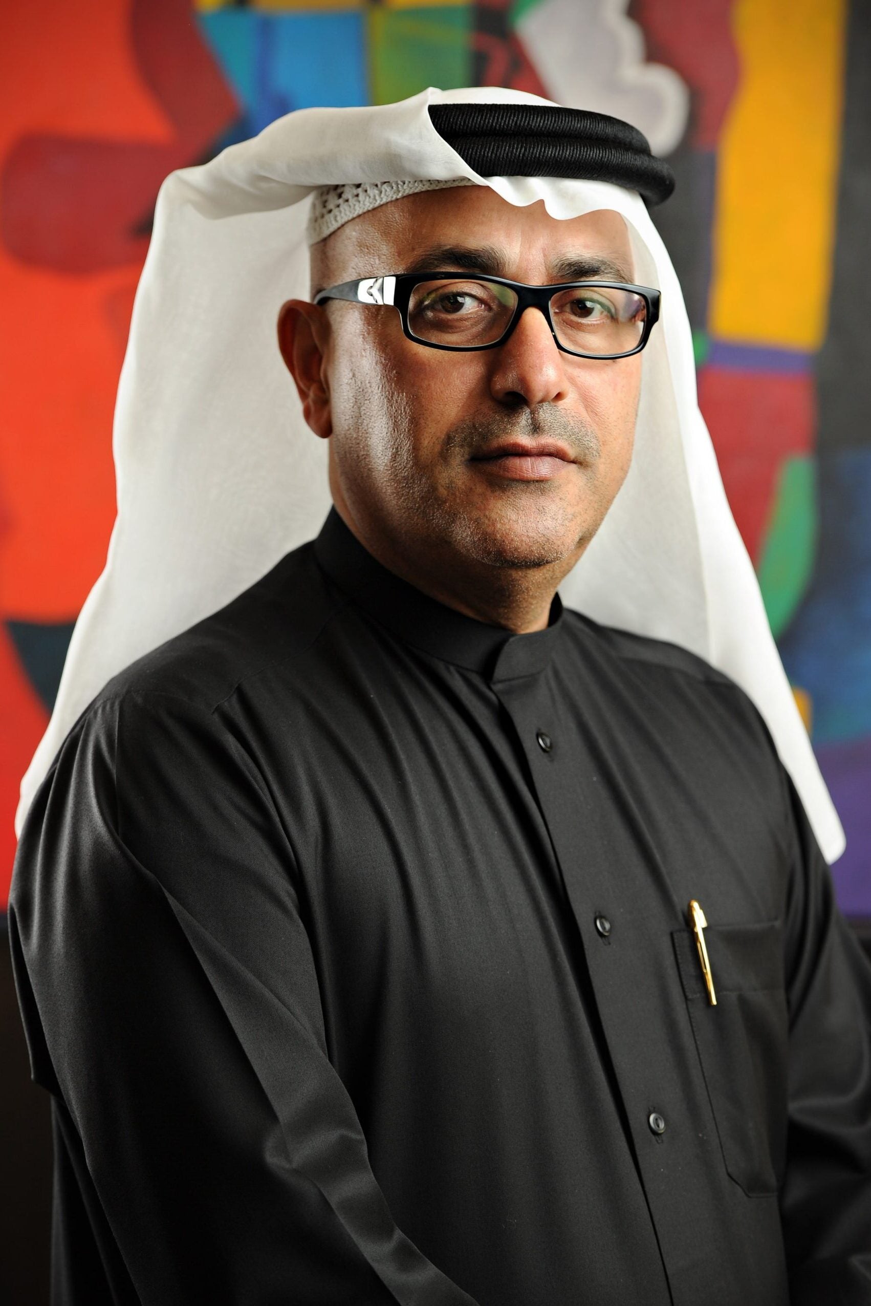 A man wearing glasses, a white keffiyeh, and a black thobe stands in front of a colorful abstract background. He has a neutral expression and a pen in his shirt pocket.
