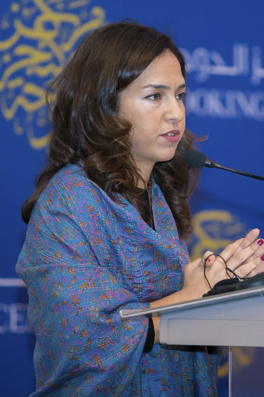 A woman with wavy brown hair speaks at a podium with a microphone, wearing a blue patterned shawl. The background features a blue display with yellow and white text and designs.