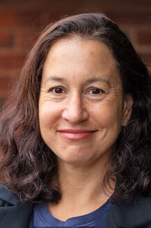 A woman with wavy dark brown hair smiles at the camera. She is wearing a dark top and stands in front of a blurred brick background.