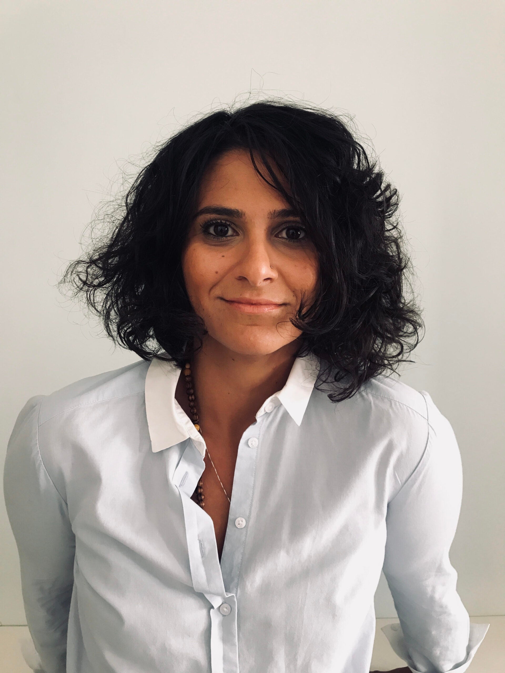 A woman with shoulder-length dark curly hair, wearing a light blue button-down shirt with a white collar, stands against a plain light background and smiles gently at the camera.