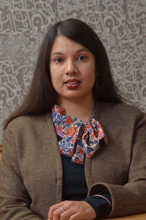 A woman with long dark hair, wearing a brown blazer and a floral scarf, sits at a table. The background features a patterned wall with intricate, abstract white designs.