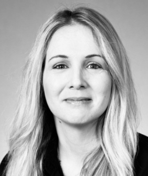 Black and white portrait of a woman with long, light-colored hair, smiling slightly and looking at the camera. She is wearing a dark top and the background is plain.