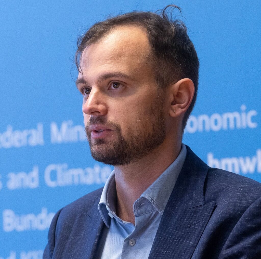 A man with short brown hair and a beard, wearing a blue suit jacket and light blue shirt, speaks in front of a blue background with partially visible white text.