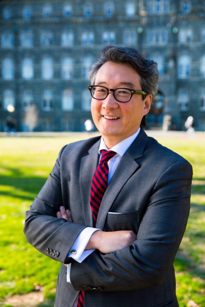 A man in a gray suit, white shirt, and red-striped tie stands outside with arms crossed, smiling. He has glasses and gray hair. A large building and green lawn are visible in the background.