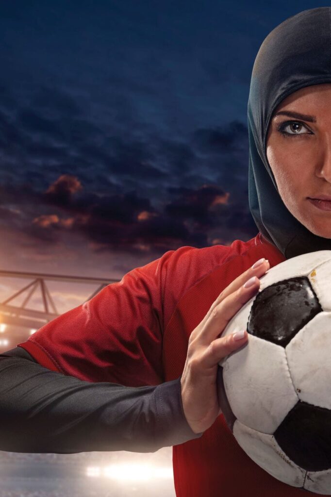 A female soccer player wearing a red jersey and a black hijab holds a soccer ball, with a dramatic stadium and evening sky in the background. Only half of her face and body are visible.