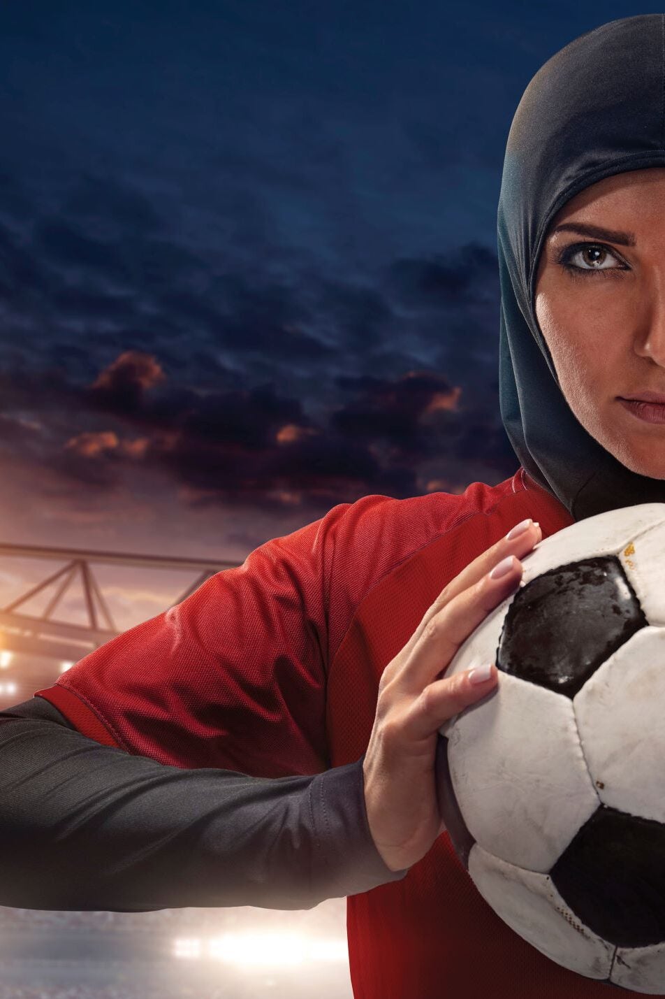 A female soccer player wearing a red jersey and a black hijab holds a soccer ball, with a dramatic stadium and evening sky in the background. Only half of her face and body are visible.