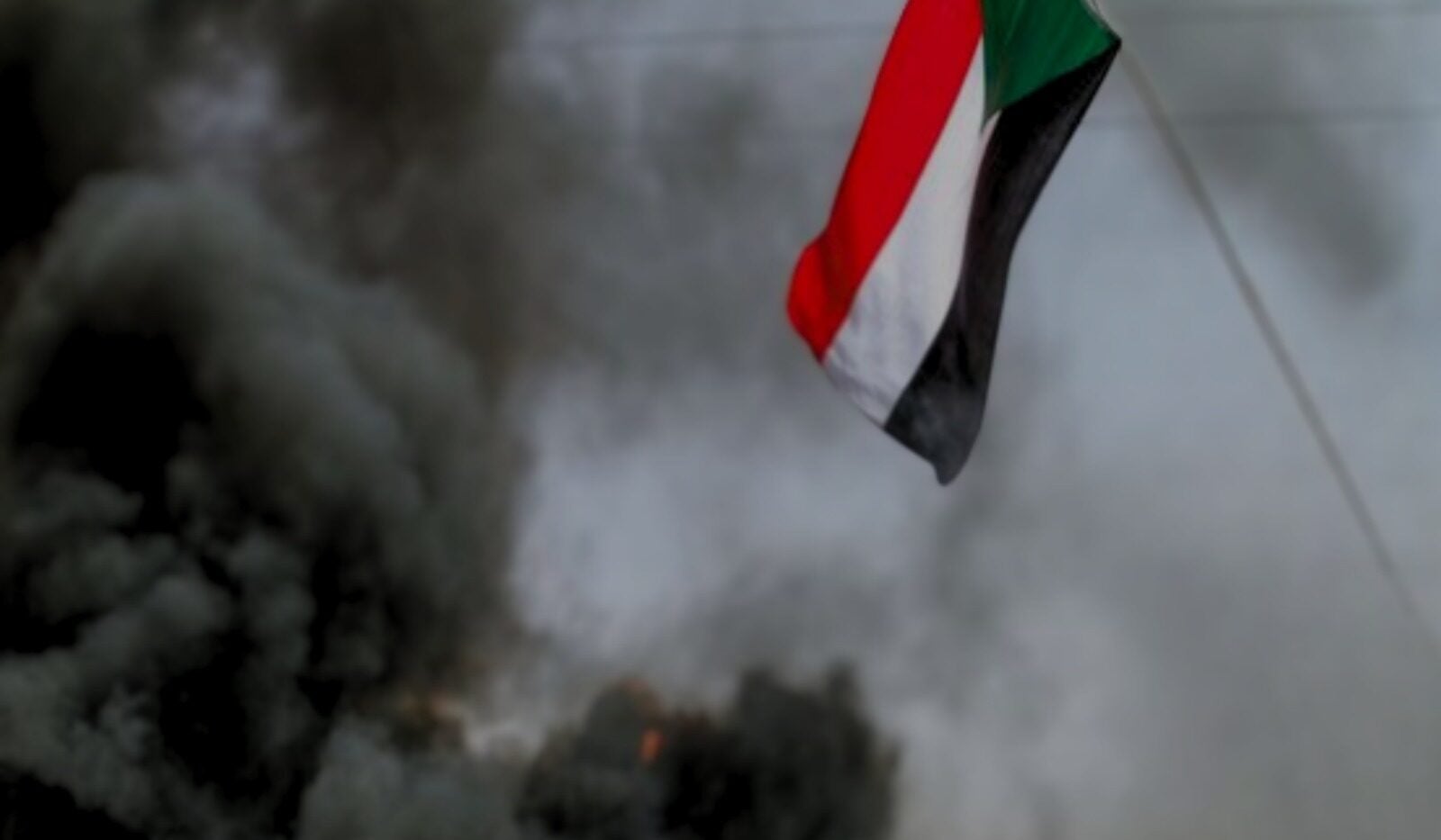A Sudanese flag waves in the air with thick black smoke and fire rising in the background, suggesting unrest or conflict.