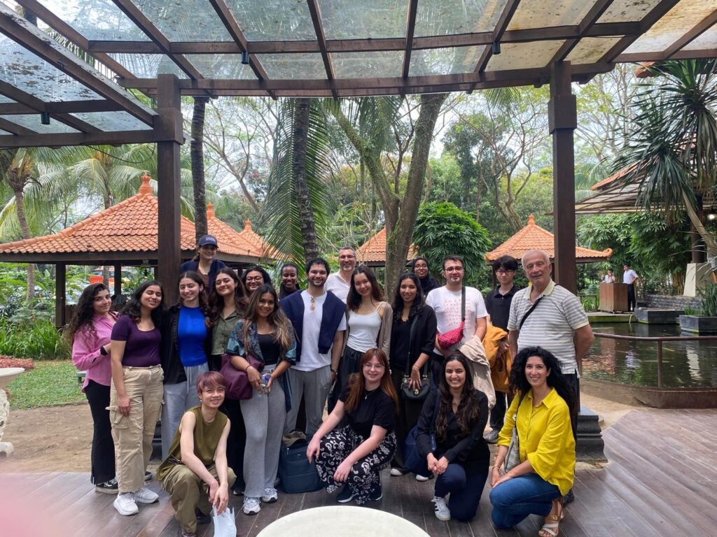 A group of people pose and smile for a photo outdoors under a glass roof, surrounded by tropical plants and trees, with red-roofed gazebos in the background.