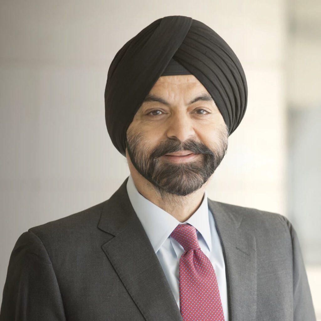 A man wearing a black turban and suit with a white shirt and red patterned tie, standing in front of a blurred light-colored background, smiling gently at the camera.