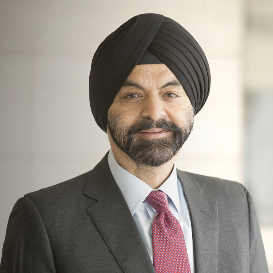 A man wearing a black turban and suit with a white shirt and red patterned tie, standing in front of a blurred light-colored background, smiling gently at the camera.
