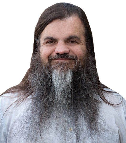 A man with long brown hair and a full, bushy beard that is darker with some gray at the center, smiling and wearing a white collared shirt, posed against a plain white background.