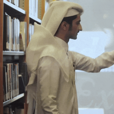 A man wearing traditional Middle Eastern attire stands by a bookshelf in a library, reaching for a book.