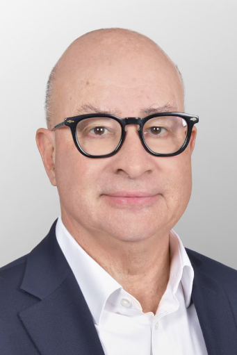 A middle-aged man with light skin, bald head, and black glasses, wearing a navy blue suit jacket and white shirt, smiles slightly against a light gray background.