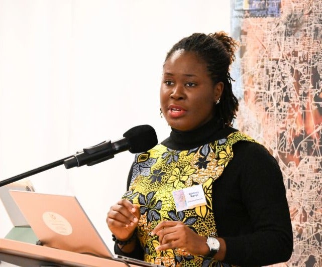 A woman wearing a black top with a colorful yellow and floral patterned dress speaks at a podium with a laptop and microphone, gesturing with her hand. A name tag is visible on her chest.