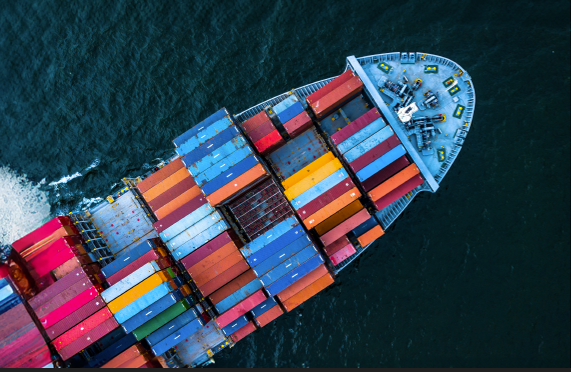 Aerial view of a large cargo ship sailing through dark blue water, carrying numerous colorful shipping containers neatly stacked on its deck.