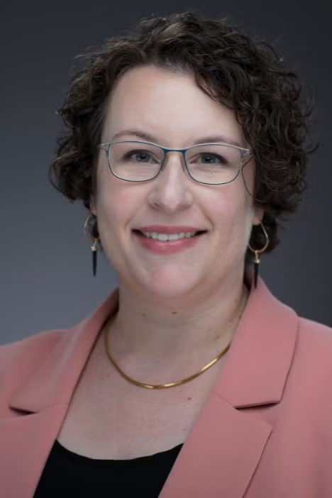 A woman with short curly hair, glasses, and earrings smiles at the camera. She is wearing a pink blazer, a light top, and a gold necklace, with a neutral gray background.