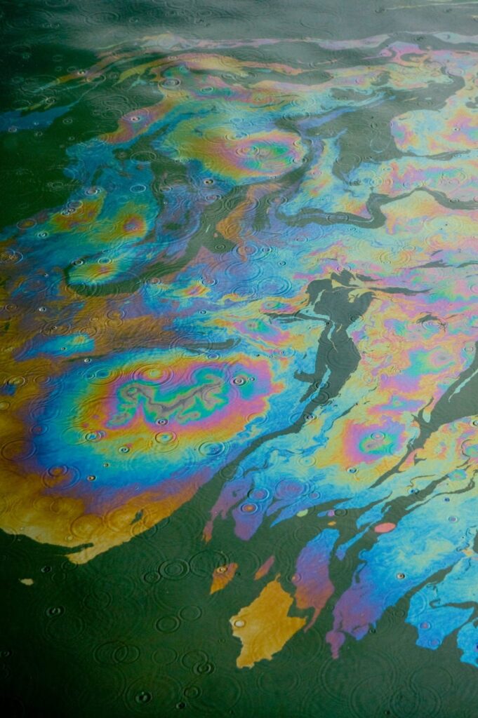 Rainbow-colored oil slick floating on green water, with swirling patterns and rain droplets creating ripple effects across the surface.