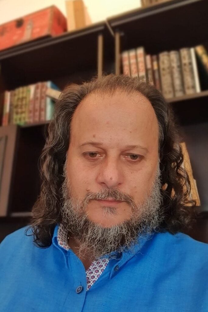 A middle-aged man with long curly hair and a gray beard, wearing a blue shirt, poses for a photo in front of bookshelves filled with books.