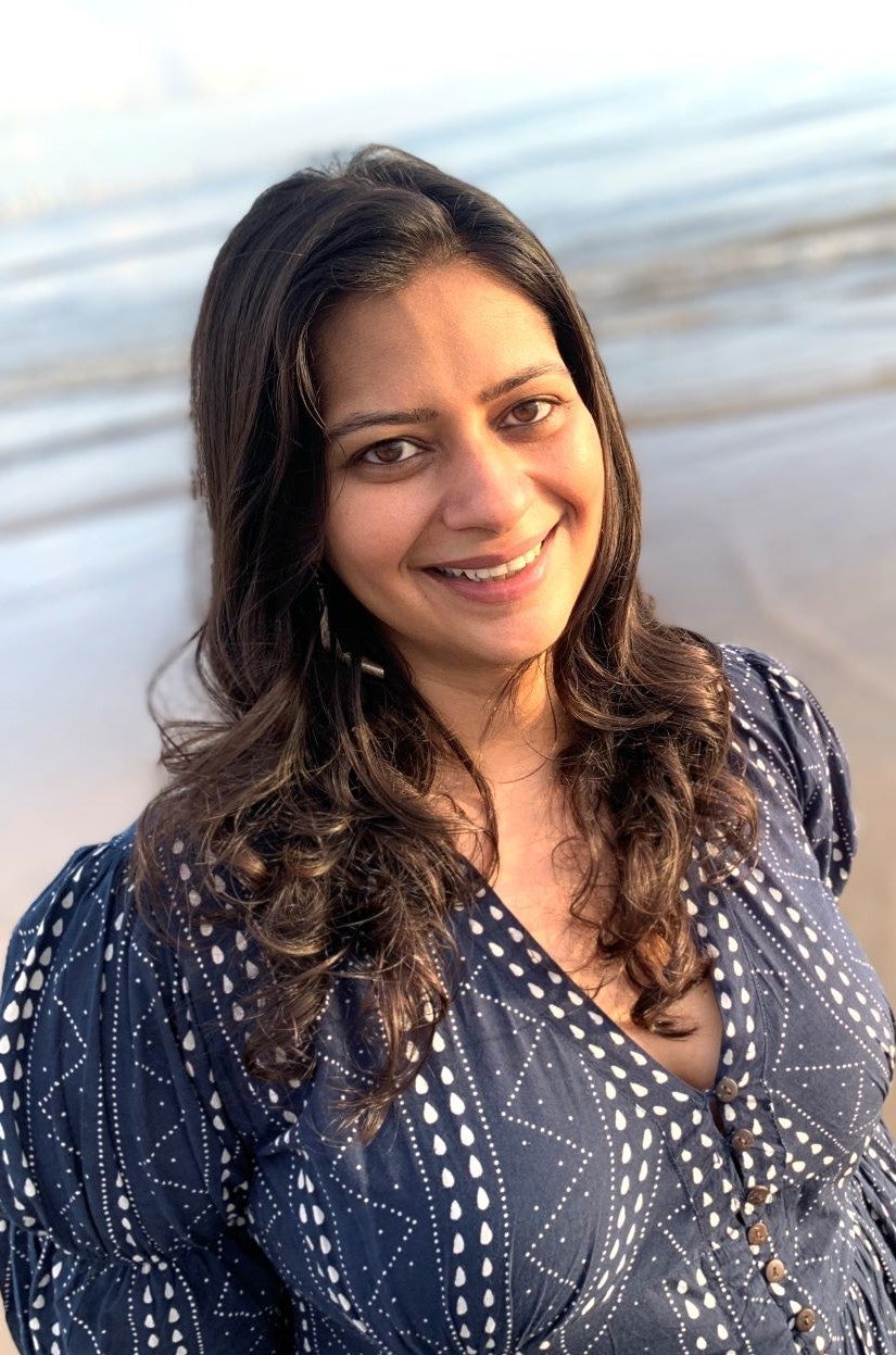 A woman with long, wavy dark hair, wearing a navy blue patterned top, smiles at the camera while standing on a beach with the ocean in the background.