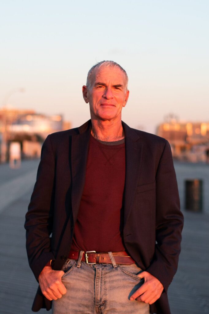 A middle-aged man with short gray hair stands outdoors in the sunlight, wearing a dark blazer over a maroon shirt and jeans, with his hands in his pockets. The background shows a blurred urban setting.