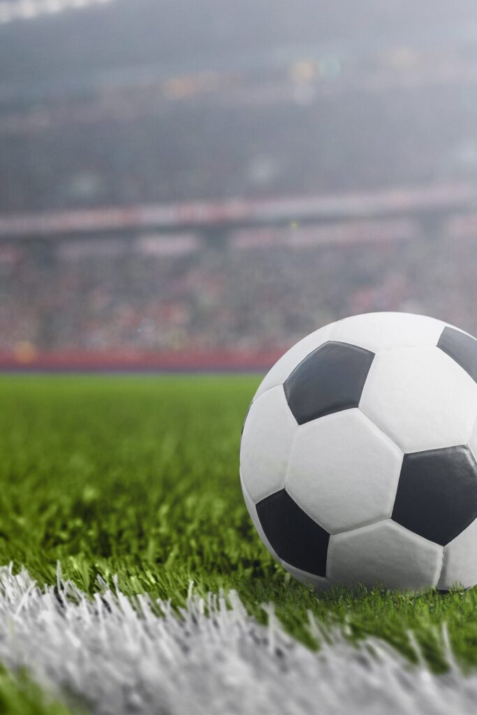A close-up of a black and white soccer ball resting on green grass near a white line, with a blurred stadium and crowd in the background.