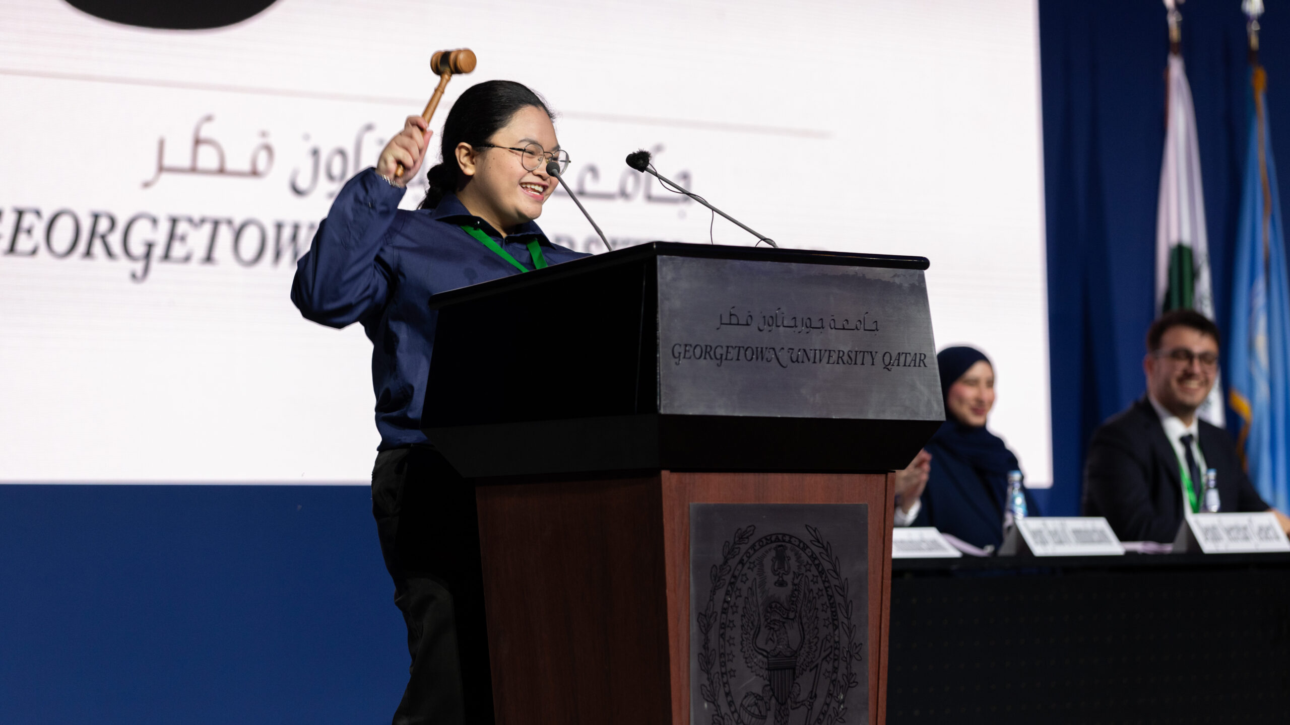 A smiling woman holds a gavel at a podium labeled Georgetown University Qatar, with three people seated at a table behind her and flags visible in the background.