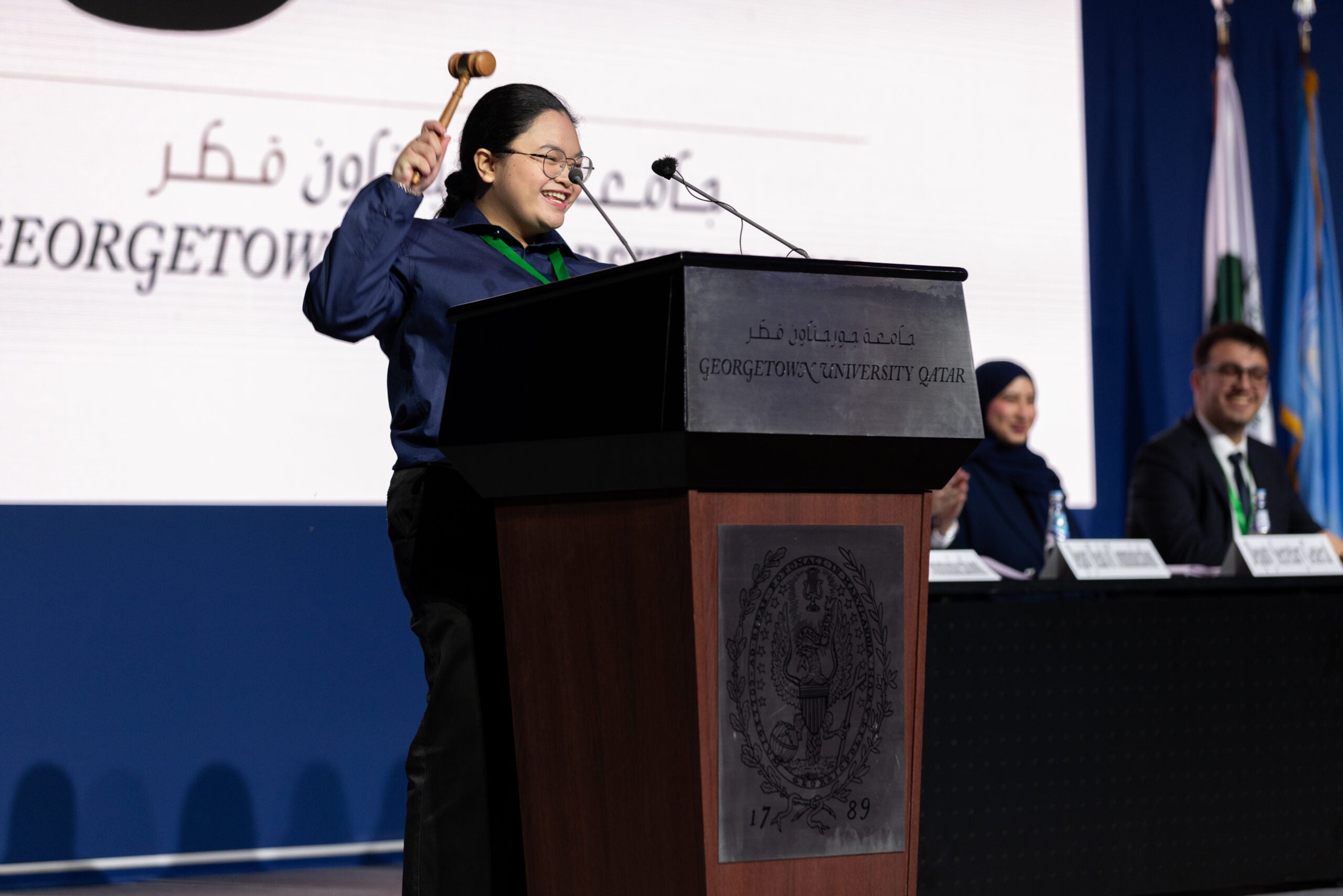 A smiling woman holds a gavel at a podium labeled Georgetown University Qatar, with three people seated at a table behind her and flags visible in the background.