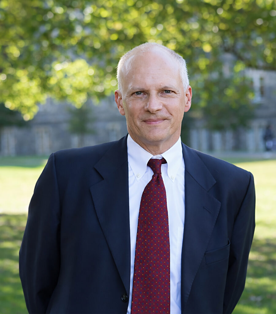 A middle-aged man with short gray hair, wearing a dark suit, white shirt, and red tie, stands outdoors in front of trees and a building, smiling slightly at the camera.