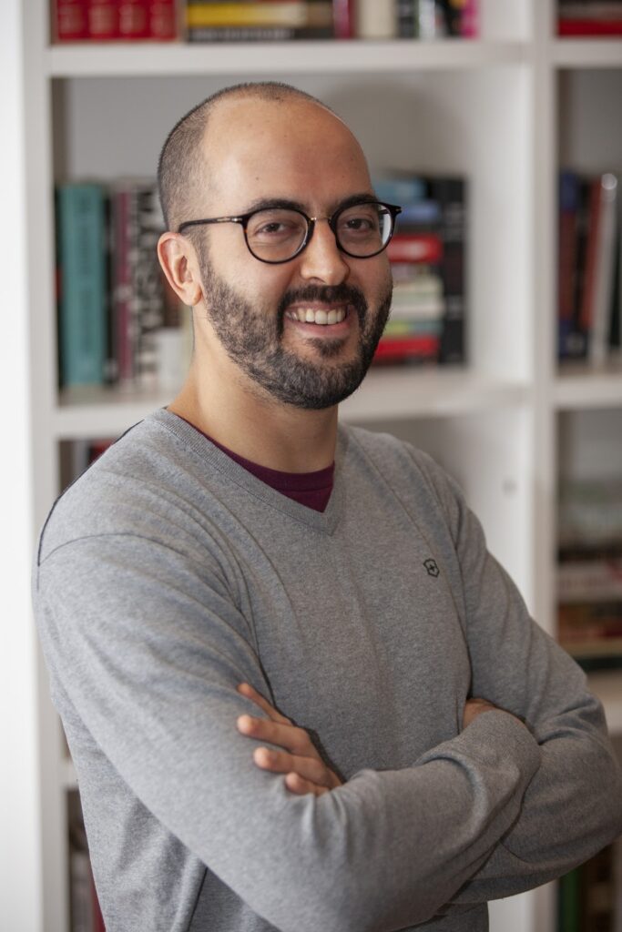 A man with short hair, a trimmed beard, and glasses stands smiling with his arms crossed. He is wearing a gray sweater and is posed in front of a white bookshelf filled with books.