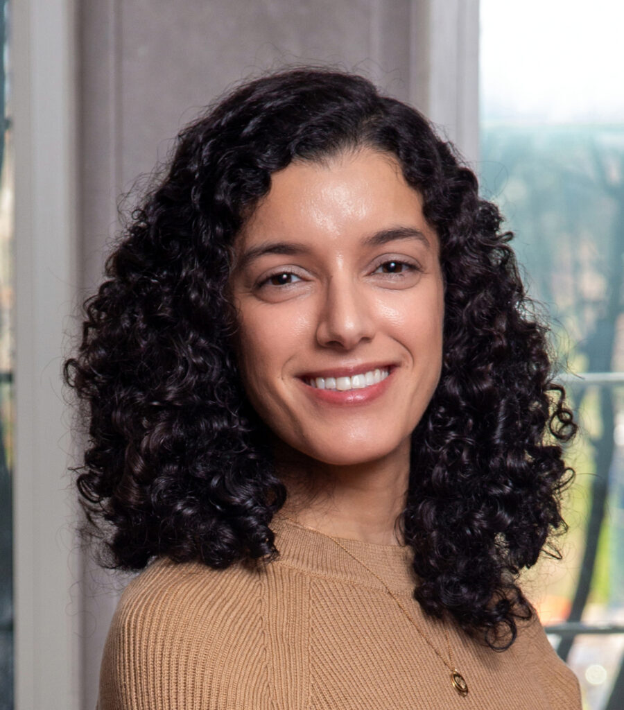 A woman with shoulder-length curly dark hair, wearing a light brown sweater and a necklace, smiles at the camera. She is indoors, with a window and trees in the background.