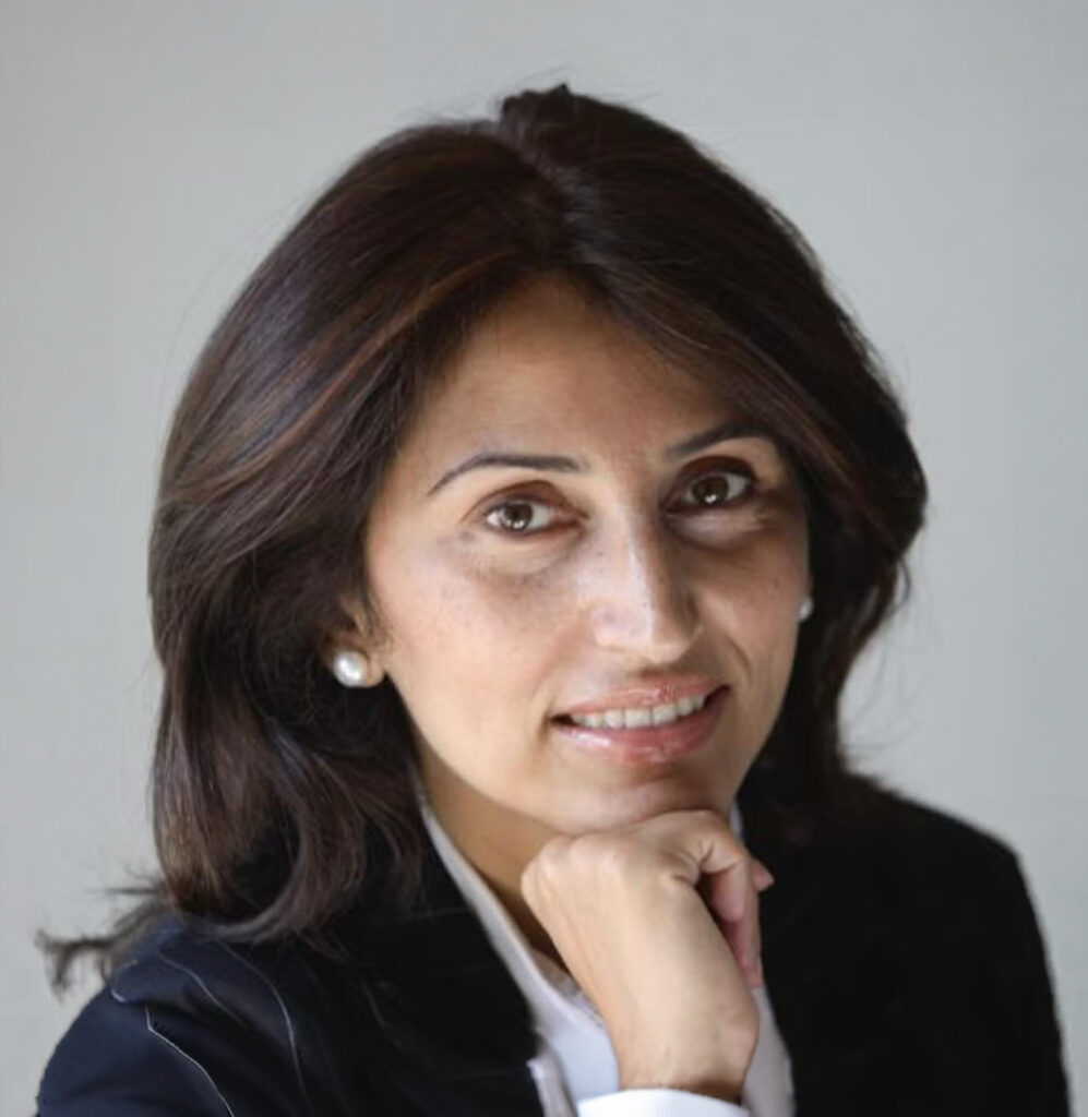 A woman with medium-length dark hair, wearing a black blazer and white shirt, rests her chin on her hand and smiles slightly at the camera against a plain light background.