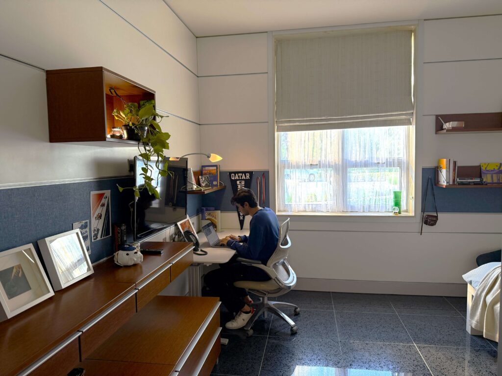 A person sits at a desk working on a laptop in a tidy, sunlit room with framed photos, shelves, plants, and a large window with blinds letting in natural light.
