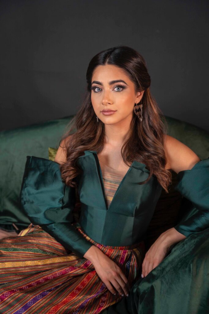 A woman with long, wavy brown hair and striking blue eye makeup sits on a green sofa. She wears an off-the-shoulder green top with puffed sleeves and a colorful, patterned skirt, looking confidently at the camera.